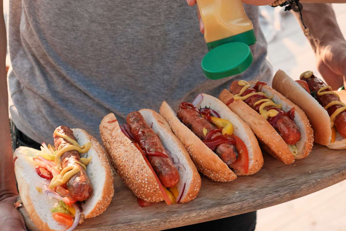 Man holds a tray of grilled hot dogs while another person adds mustard