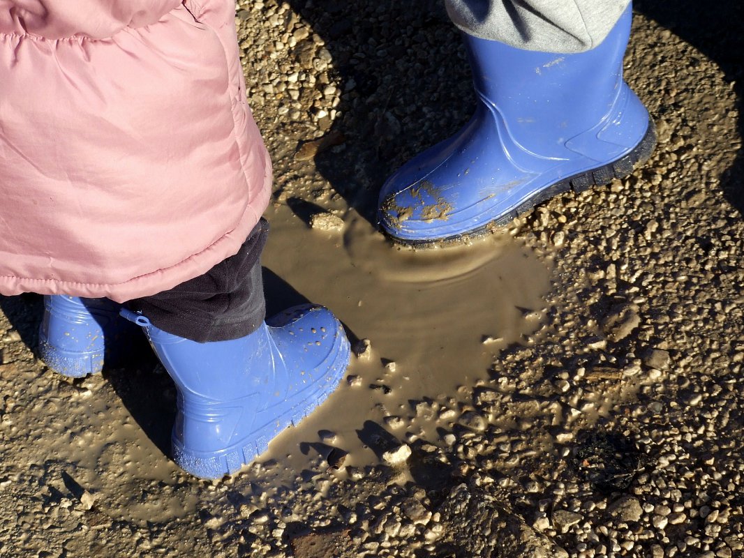 Two kids playing in a mud puddle in blue gumboots