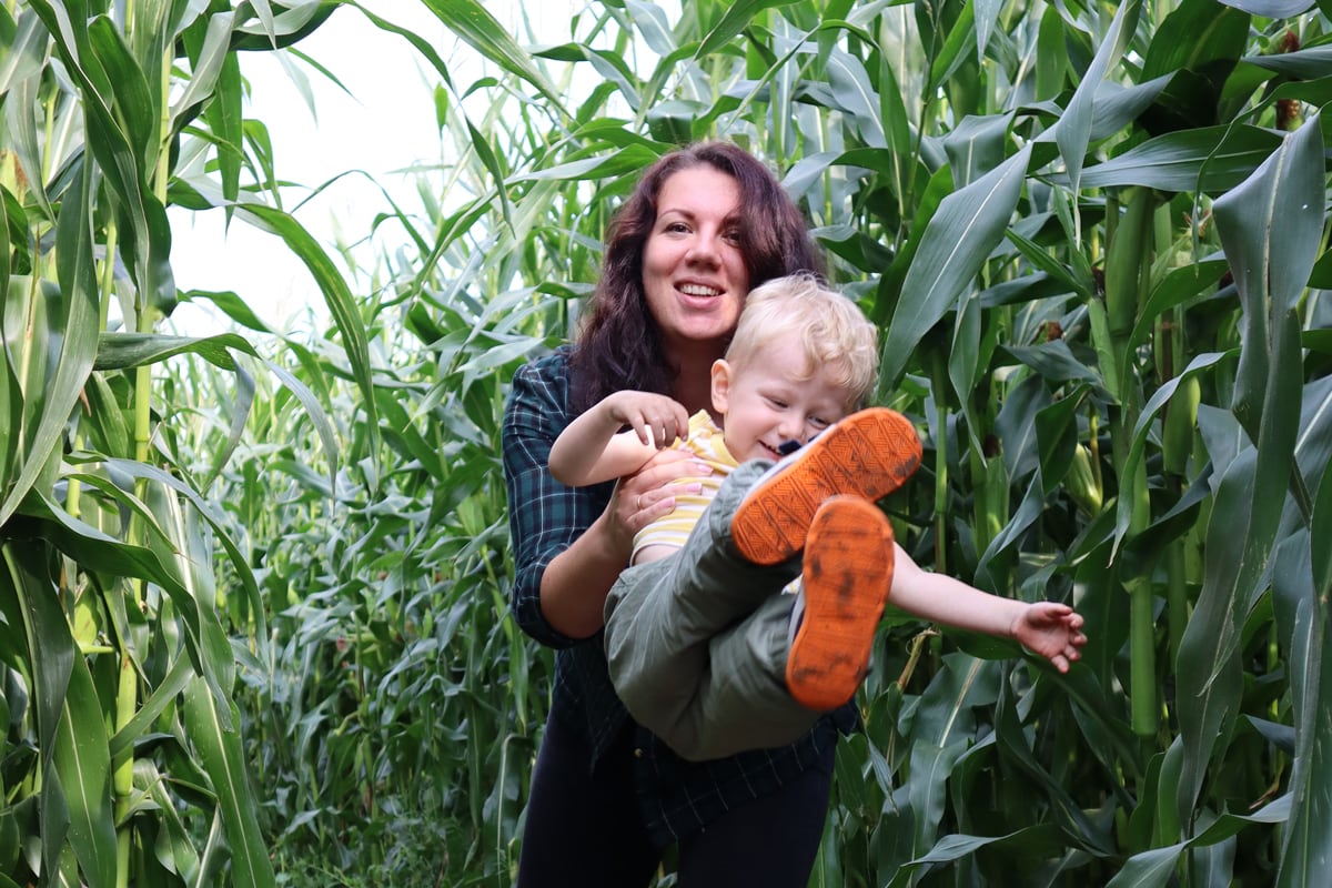A mother swings a young boy through the air while walking through a path in a dense field of corn