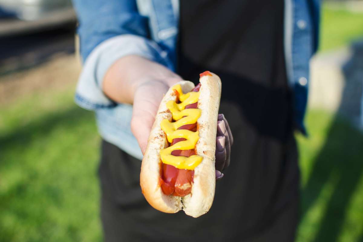 Woman holding a hot dog with plenty of mustard on it towards the camera